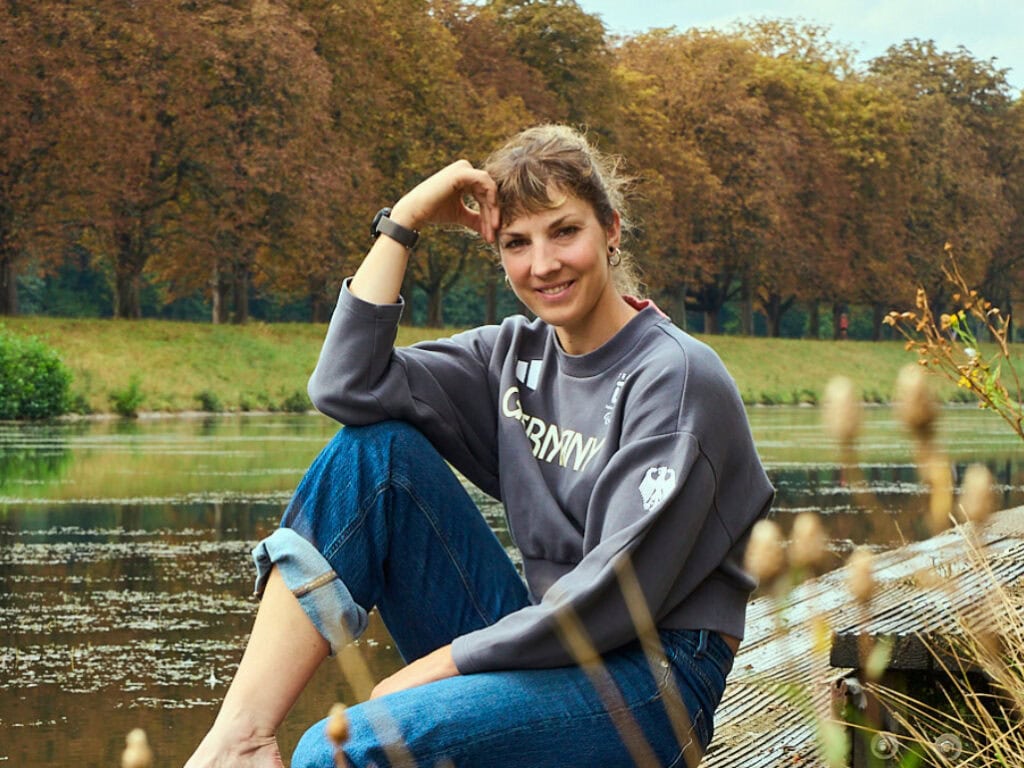 Ruderin Kathrin Marchand (Blonde Haare, Blue Jeans, Pulli) sitzt entspannt auf einem Steg am Decksteiner Weiher in Köln Lindenthal.